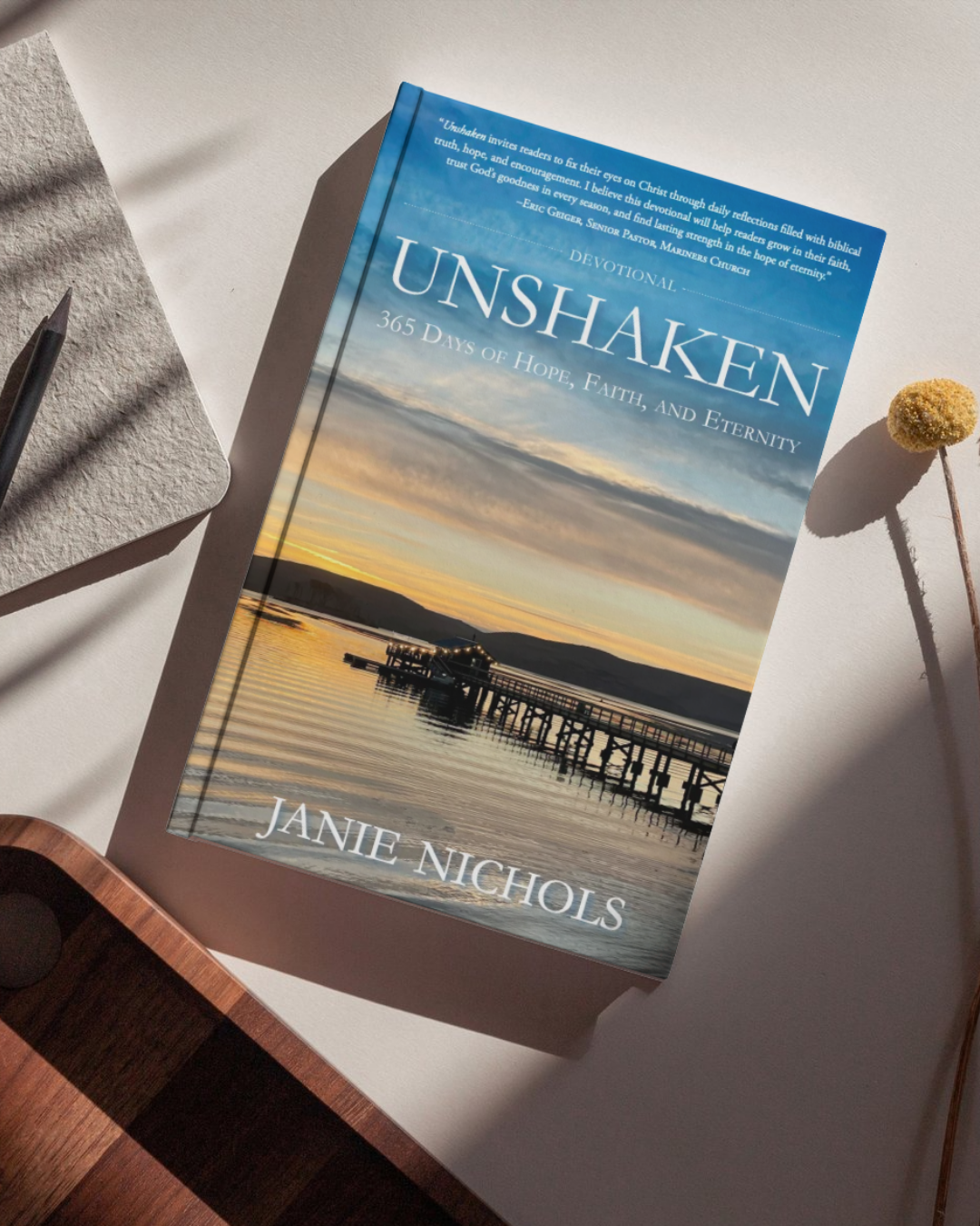 The book 'Unshaken' by Janie Nichols is displayed diagonally on a light beige surface with artistic shadows. The cover features a scenic photograph of a sunset over water with a pier, and the title is written in large white letters.