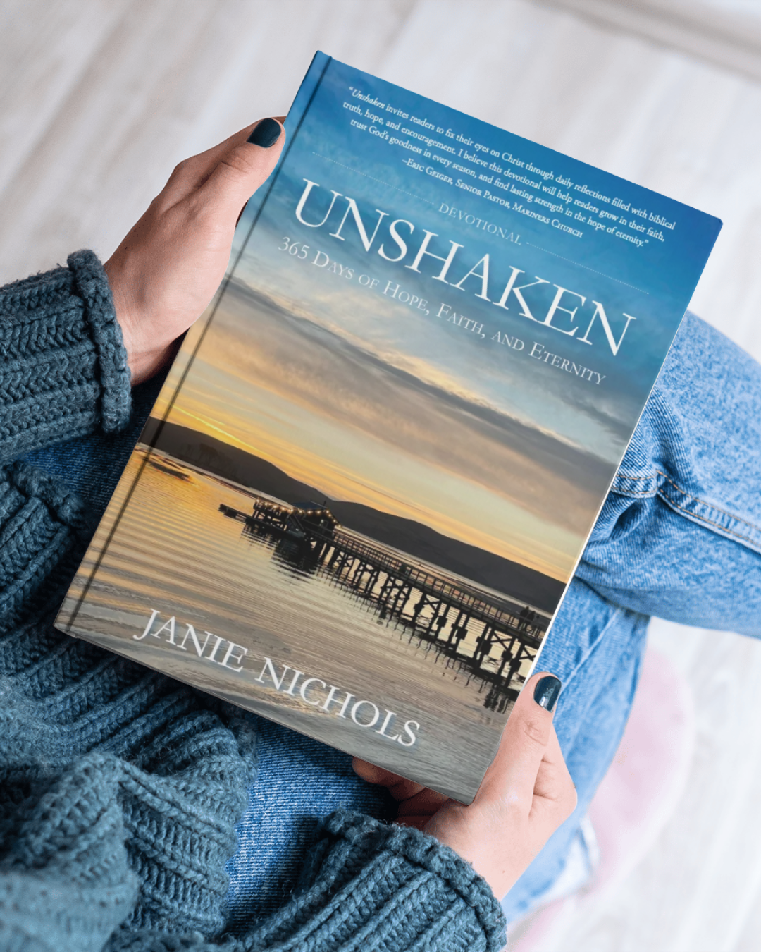 A person holding a hardcover book titled "Unshaken" by Janie Nichols. The book cover shows a scenic sunset over water with a pier and mountains.